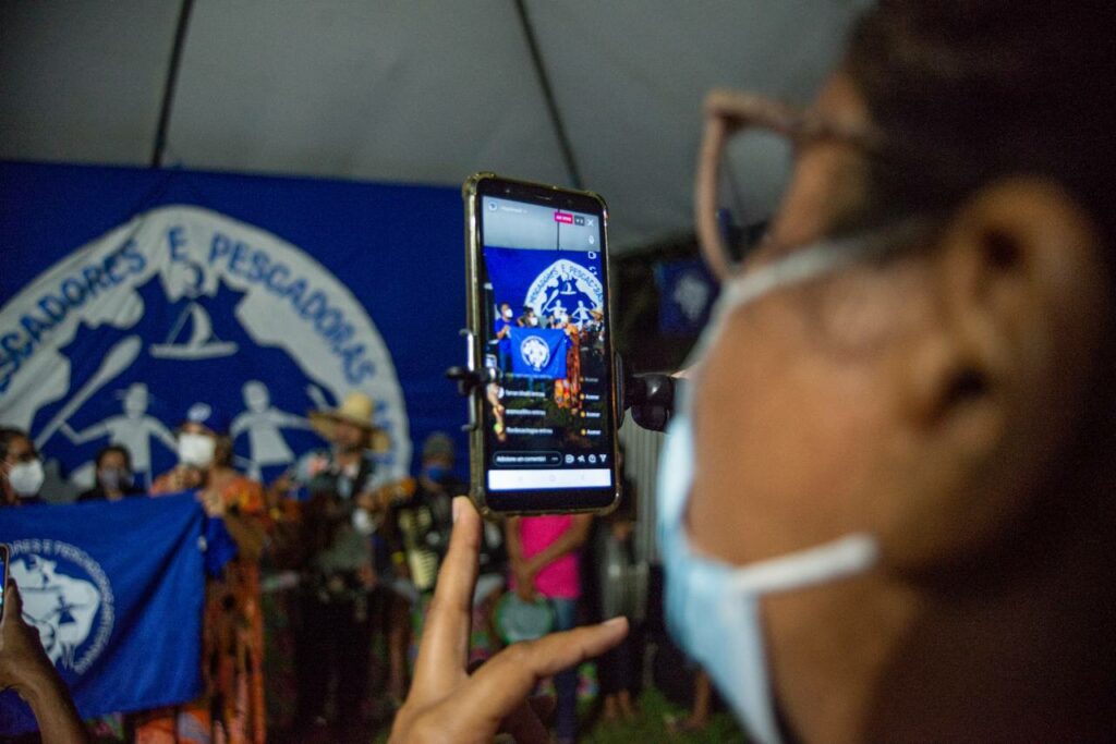 Mulher segura celular em transmissão ao vivo da abertura do evento Grito da Pesca. A foto mostra seu rosto e o celular diante de três pessoas que seguram a bandeira azul do MPP. Uma delas fala ao microfone