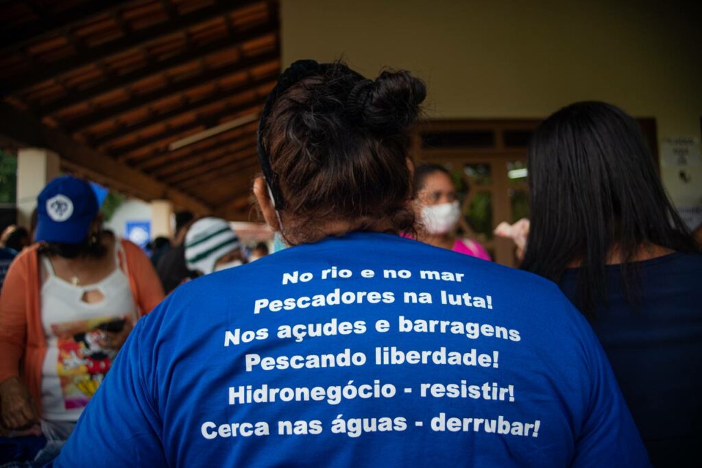 Foto de uma mulher de costas num salão. Ela usa cabelo preso em coque e blusa azul onde se lê a seguinte poesia: "No rio e no mar / Pescadores na luta! / Nos açudes e barragens / Pescando liberdade / Hidronegócio: resistir! / Cerca nas águas: derrubar!"